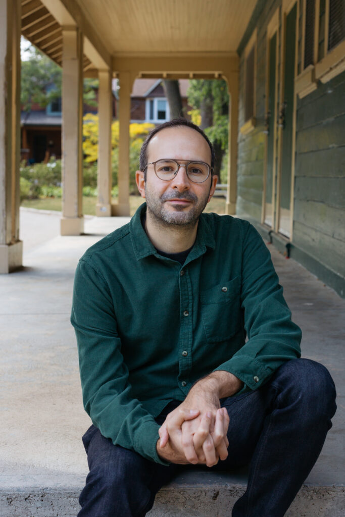 Babak Lakghomi sits on a veranda with his hands clasped together. He is wearing a forest green shirt and glasses with big round frames.