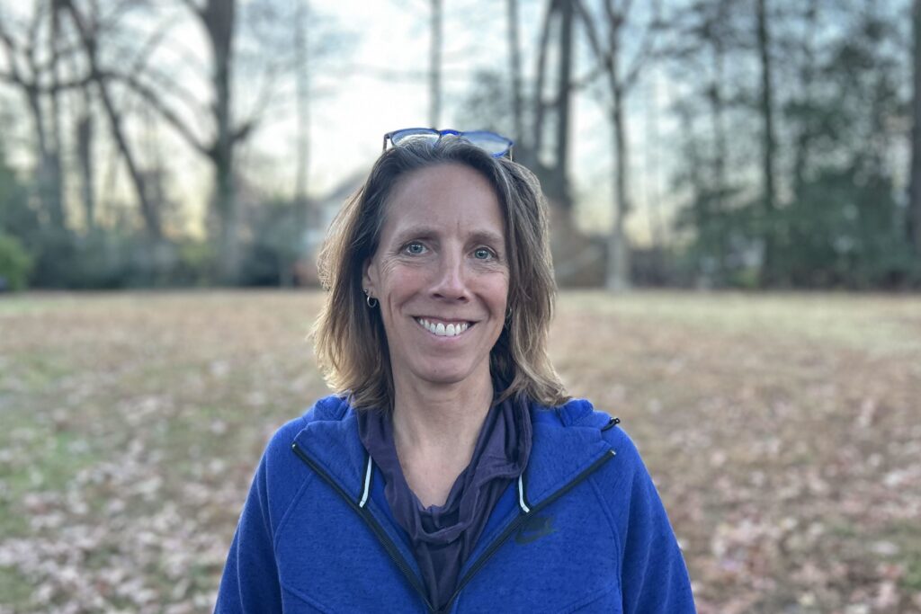 writer Deb Werrlein, a white woman standing in a natural, wooded area, smiling and wearing a blue hoodie