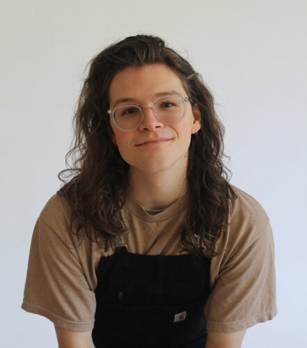 CD Eskilson, a trans-nonbinary person with long brown hair, clear-framed glasses, and black overalls over a tan shirt. They are smiling and have thin hoop earrings