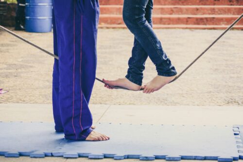 two people engaging in tightrope walk practice from the knees down, one person is standing and the other is balanced on a tightrope positioned not far from the ground with a safety mat