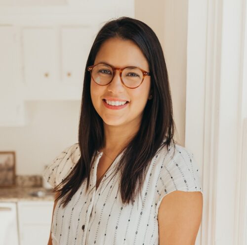 Cristi Donoso, an Ecuadorian American woman, standing in a doorframe and wearing a button down white shirt with some vertical stripes. She's wearing brown glasses and smiling, and a home's cabinet and counter are blurred behind her.