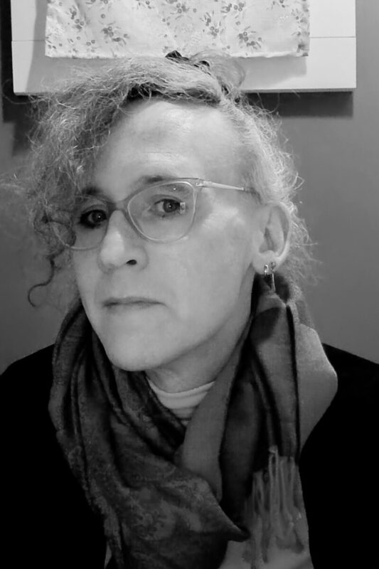 Cassandra Whitaker looks at the camera, her head turned at a slight angle. They are wearing glasses with large oval frames, and a fringed scarf.